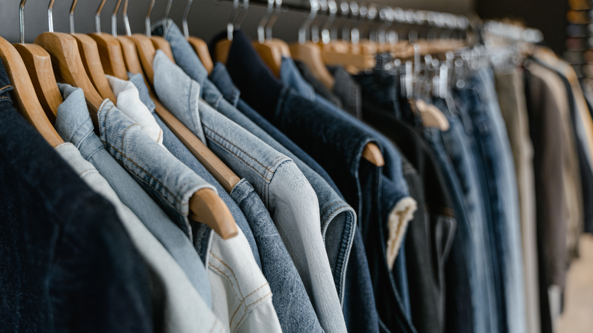 A row of denim jackets and shirts hanging on wooden hangers on a clothing rack.