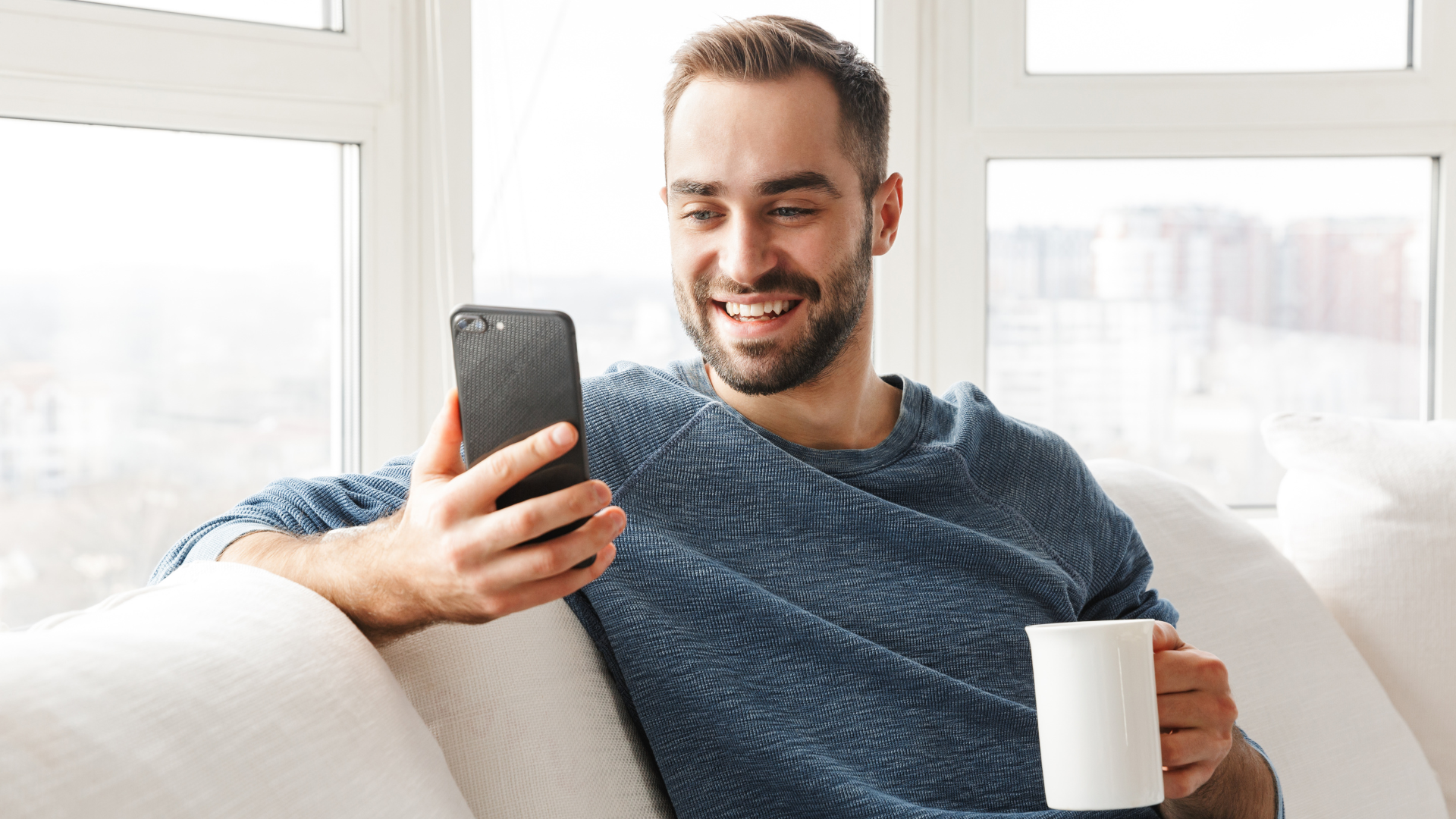 A man in a blue sweater sits on a couch, holding a smartphone in one hand and a white mug in the other, smiling in a brightly lit room with large windows.