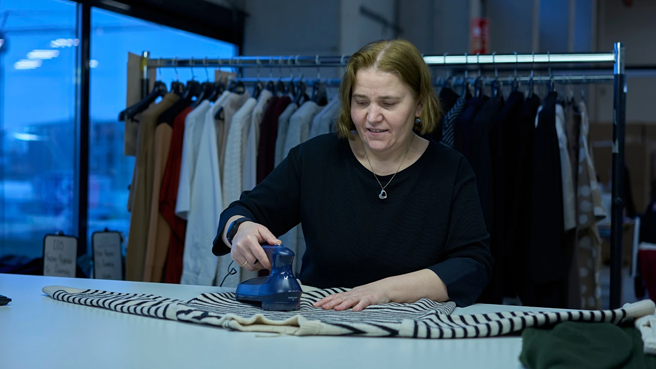 A person ironing a striped garment on a table in a clothing store, with a rack of hanging clothes in the background.