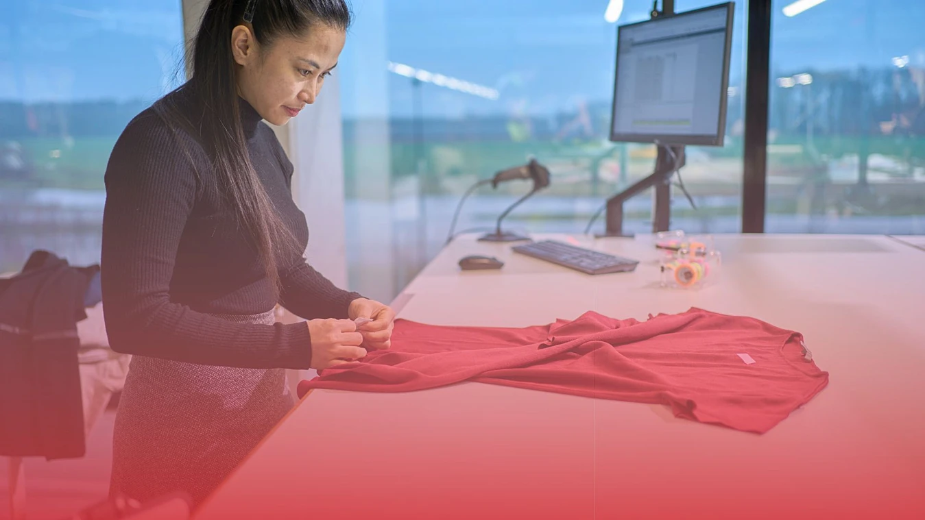 A woman stands at a desk examining a red garment in a well-lit workspace, with a computer and the view of a window in the background.