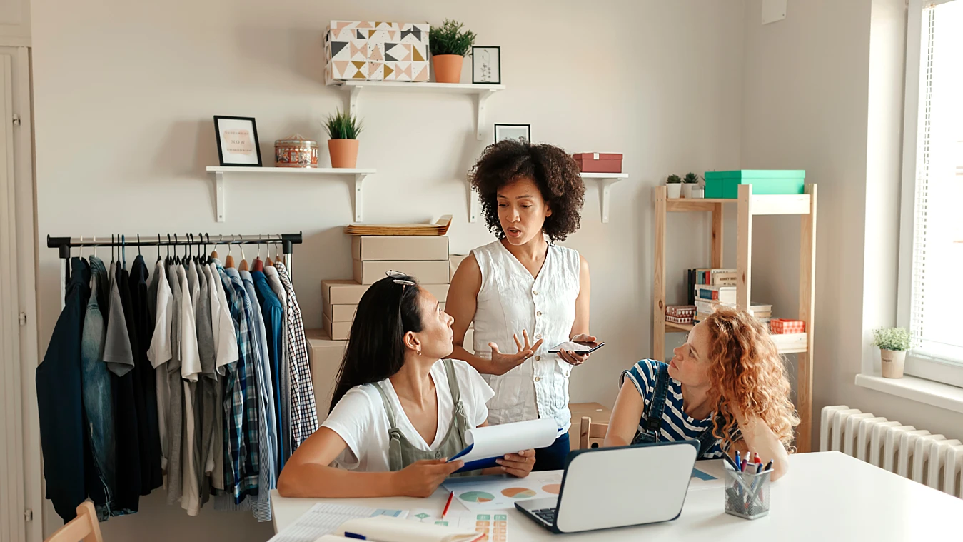 Three women are having a discussion in a room with clothing racks and shelves. One stands holding a phone, while the other two sit at a table with a laptop and papers.