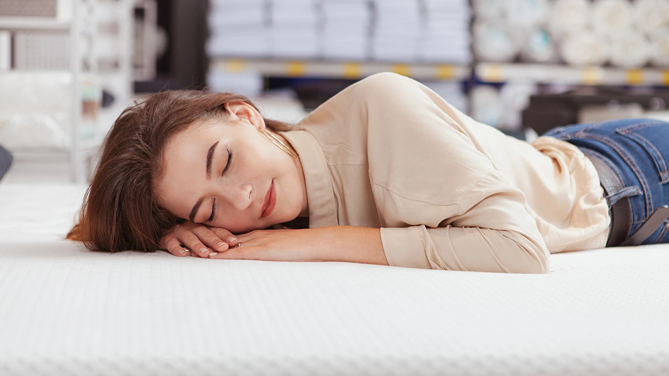 Woman lying on a mattress in a store, eyes closed and smiling, with shelves of stacked items in the background.