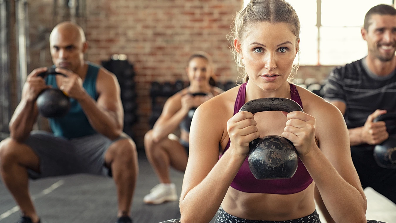 People exercising with kettlebells in a gym, focusing on a woman in a purple sports bra.