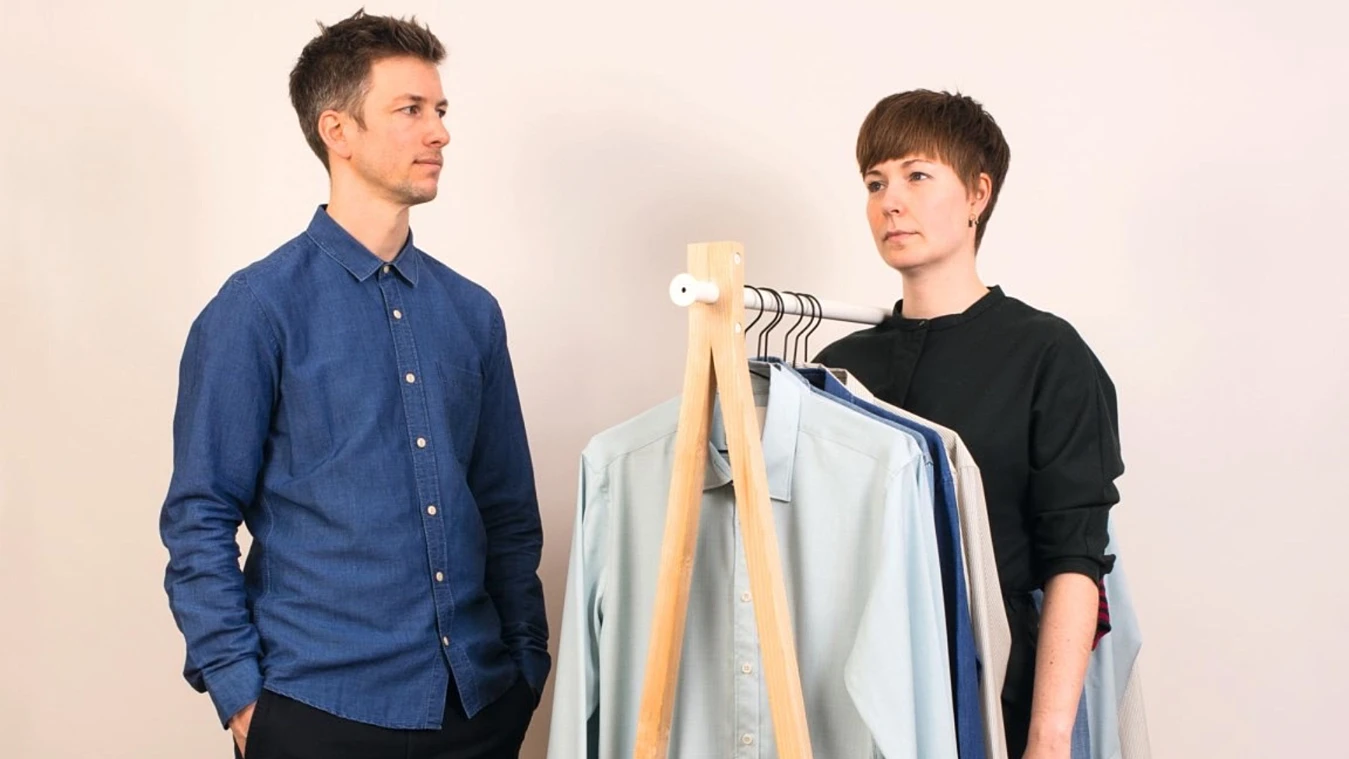 Man and woman standing by a clothes rack with various shirts on hangers, looking at each other expressionlessly.