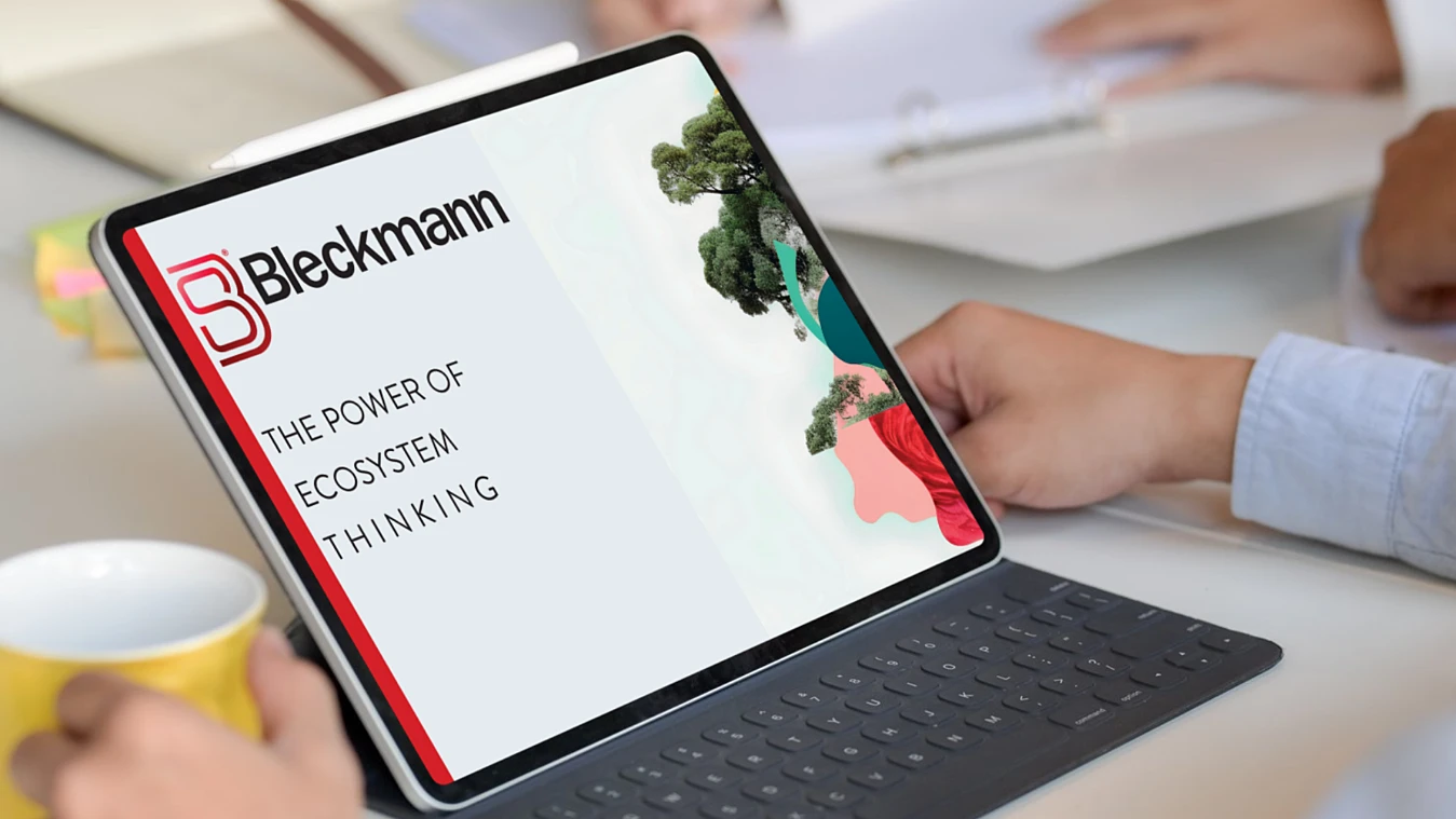 A person holding a tablet displaying a presentation titled "The Power of Ecosystem Thinking" with the Bleckmann logo. A keyboard, cup, and more hands are visible in the background.