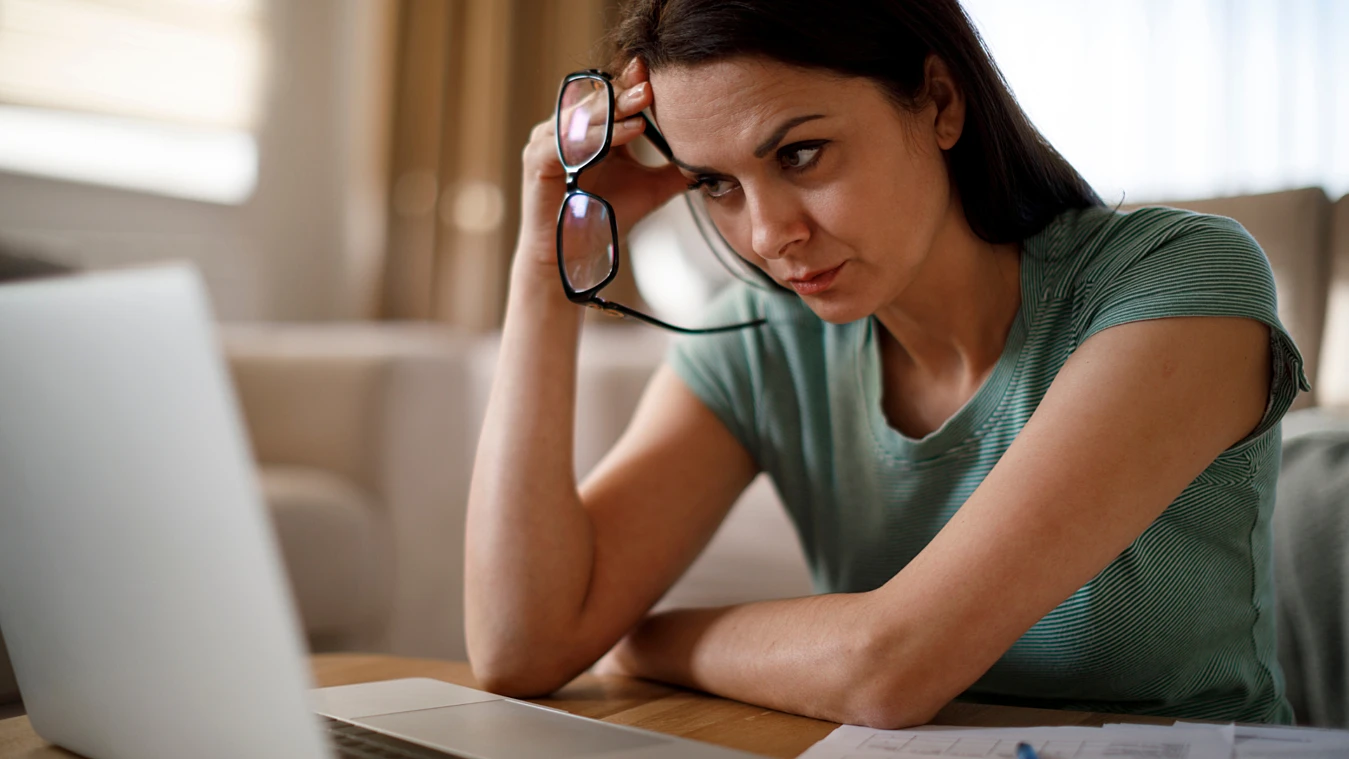 Woman in a green shirt looks intently at a laptop screen, holding her glasses in one hand, with papers on the table.