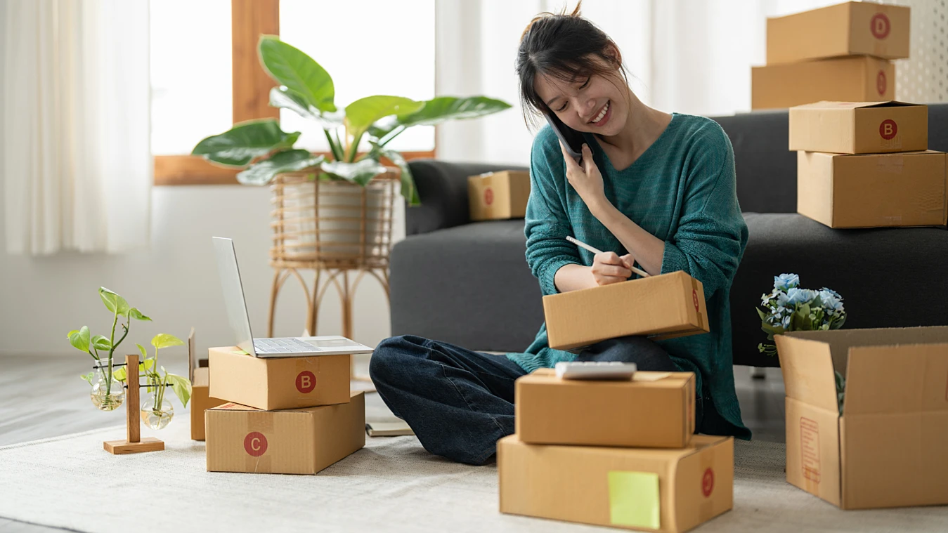 A woman sits on the floor, talking on the phone and writing on a package. She is surrounded by boxes, a laptop, and a plant in a well-lit room.