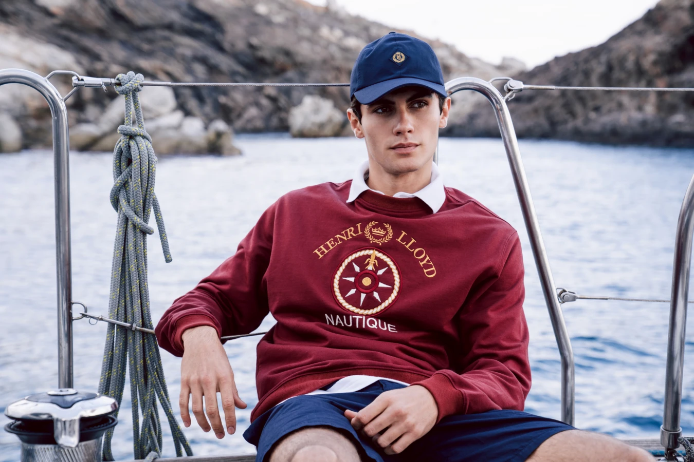 Person wearing a red sweatshirt and blue cap, sitting on a boat by rocky water's edge.