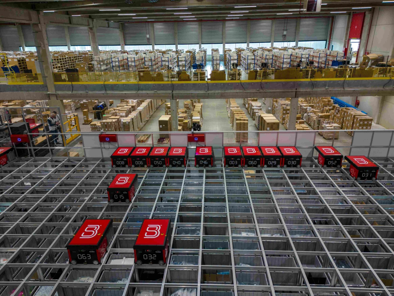 A warehouse interior with numerous shelves and boxes. Red robotic units labeled "B" move across a grid on the floor, likely for automated sorting or storage.