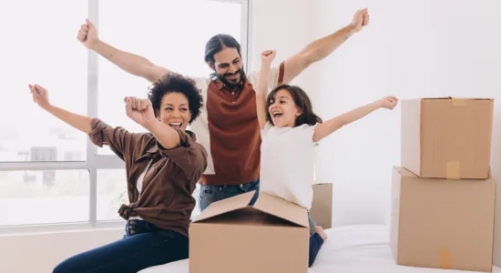 A happy family of three celebrates on a bed surrounded by packed boxes, suggesting a recent move or new home.