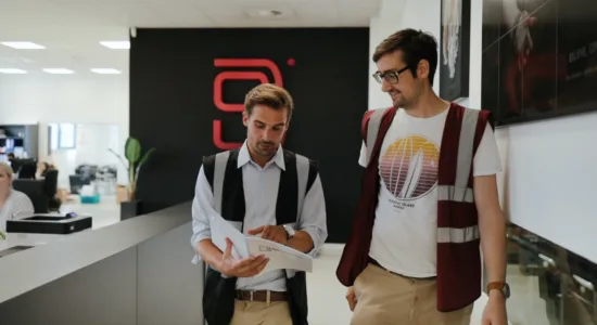 Two men wearing vests walk through a modern office, discussing documents. One holds papers, and the other wears glasses and a t-shirt. A logo is visible on a black wall in the background.