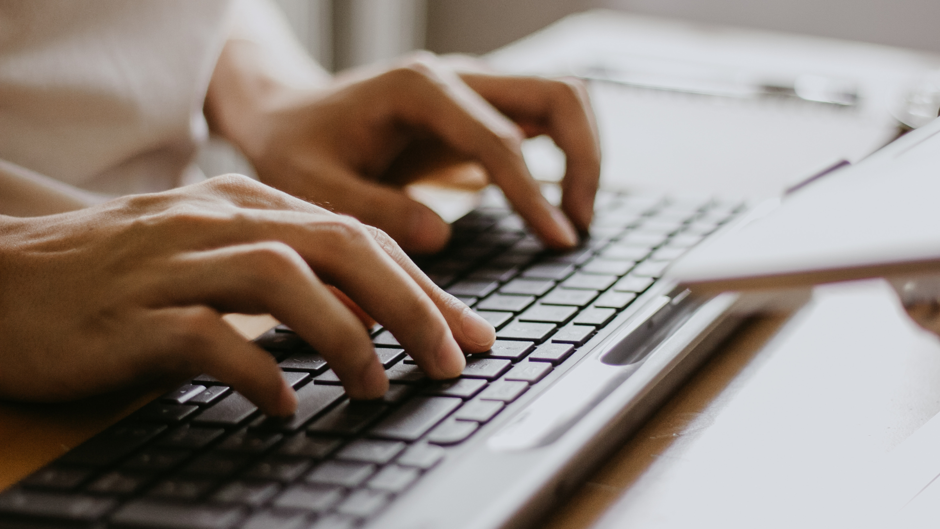 Hands typing on a black keyboard with a notepad beside it.