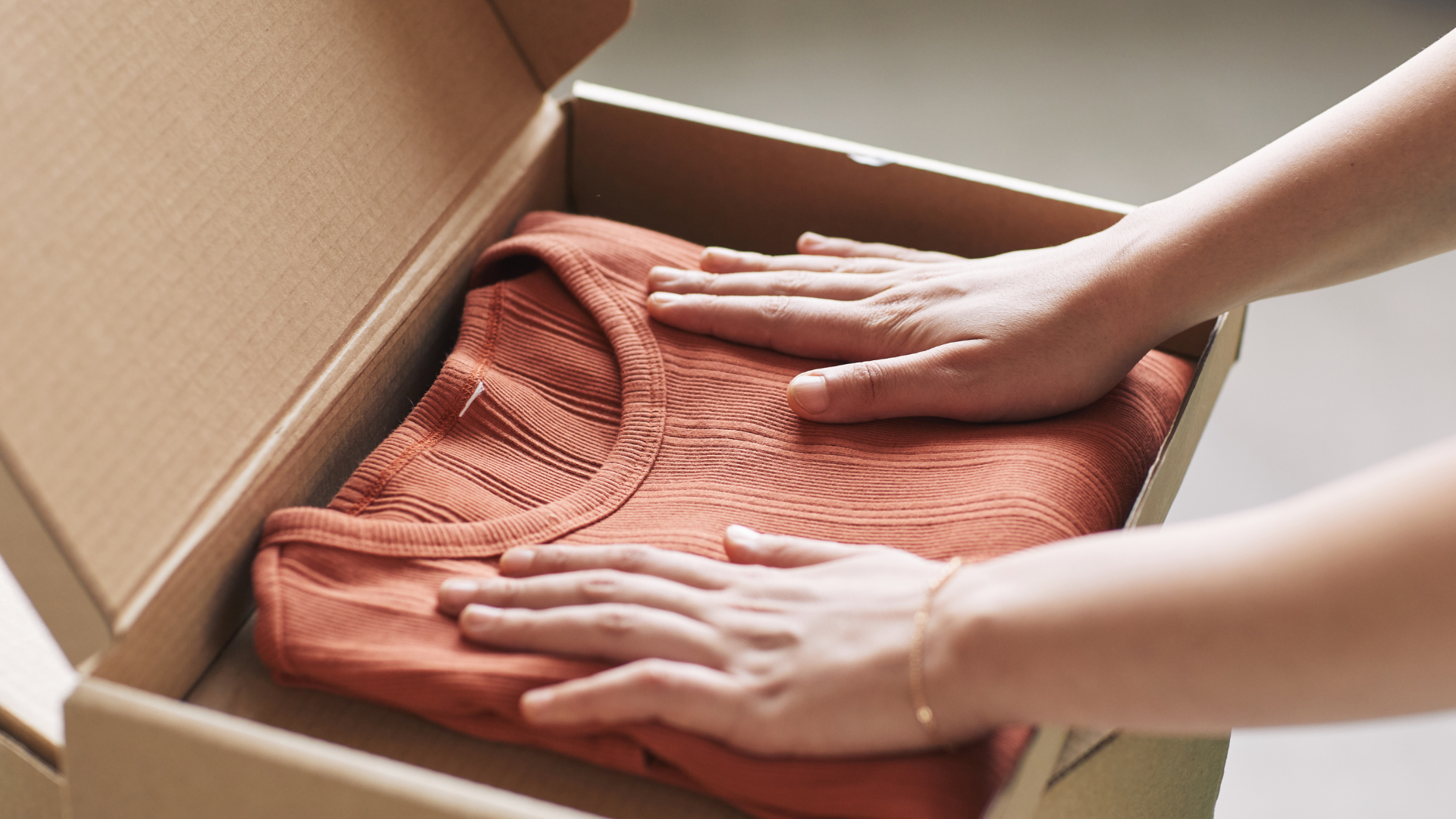 A folded orange shirt placed in an open cardboard box, with two hands gently packing it.