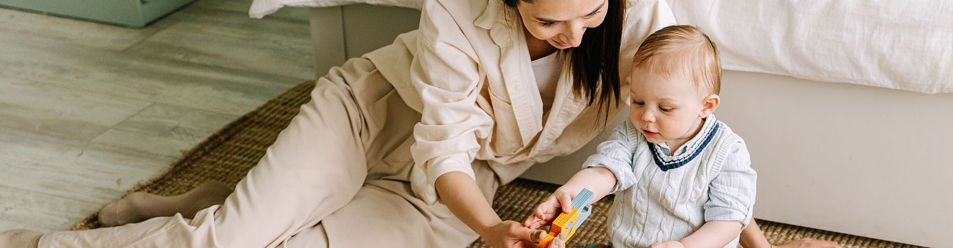 Adult and toddler sitting on the floor, playing with blocks beside a bed.