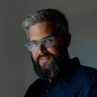 A man with gray hair, a beard, and glasses wearing a dark shirt stands against a gray background.