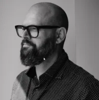 Black and white image of a man with glasses and a beard, looking to the side, wearing a dotted shirt.