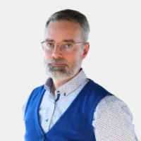 A man with a gray beard and glasses, wearing a blue vest and patterned shirt, stands against a plain white background.