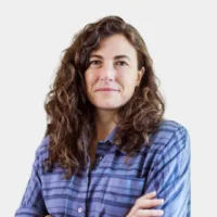 Person with long curly hair wearing a blue striped shirt, standing against a plain background.