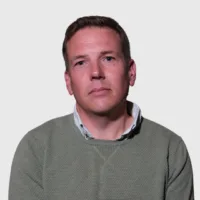 A person wearing a green sweater and white shirt is looking directly at the camera against a plain white background.